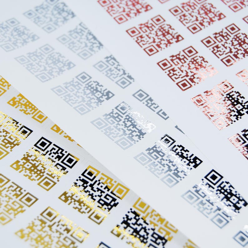 May include: Sheets of QR code stickers in various metallic colors, including gold, silver, and copper, arranged diagonally. The codes are printed on a white background, creating a contrast between the shiny codes and the matte paper.