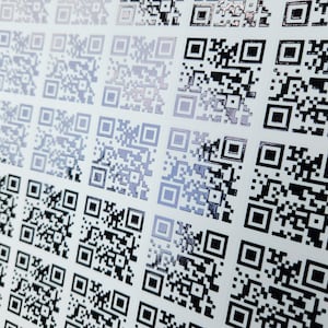 May include: Close-up of a sheet of white paper covered in rows of black and silver QR codes. The codes are square with a mix of black and silver squares and rectangles. The codes are arranged in a grid pattern.
