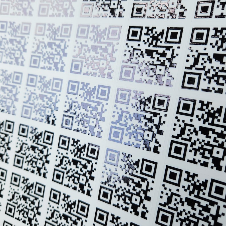 May include: Close-up of a sheet of white paper covered in rows of black and silver QR codes. The codes are square with a mix of black and silver squares and rectangles. The codes are arranged in a grid pattern.