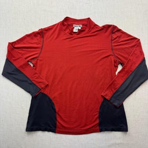 May include: A long-sleeve mock neck shirt in red with dark navy blue side panels and sleeve undersides. The garment features a WinterSilks brand label at the inner neckline and visible dark stitching along the seams of the sleeves and torso.
