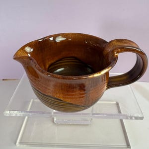 May include: A ceramic creamer pitcher featuring a glossy, variegated brown glaze with a smooth finish. The vessel has a wide, rounded body with horizontal ribbed detailing around the base, a curved pouring spout, and a sturdy, arched handle.