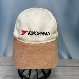 May include: A baseball cap with an off-white crown and a tan suede-like brim. The front of the crown features the red Yokohama logo with a stylized Y symbol followed by the black text YOKOHAMA embroidered in a bold, sans-serif typeface.