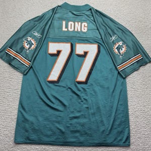 Pode incluir: Camisola de futebol turquesa com o nome "LONG" e o número "77" nas costas. A camisola tem detalhes em laranja e branco, com o logotipo dos Miami Dolphins nas mangas. A camisola é feita de material de malha respirável.
