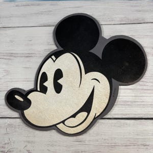 May include: A flat, die-cut wall decal or sign featuring the classic head silhouette of Mickey Mouse. The design displays a black outline and ears, with a cream-colored face, black nose, and a wide, smiling mouth. The word Disney is printed on the side.