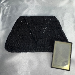May include: A black beaded clutch bag featuring a V-shaped pattern of elongated beads on the front surface. Beside the clutch is a rectangular, gold-toned metal frame containing a faded, aged photograph with visible wear and discoloration.