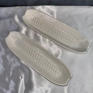 May include: Two elongated, off-white ceramic serving dishes shaped like corn cobs. Each dish features a textured surface mimicking the appearance of individual corn kernels arranged in rows, with smooth, curved ends and a glossy, reflective finish.