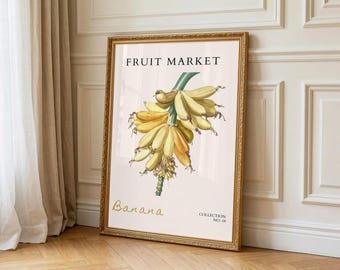 May include: A framed print with a botanical illustration of bananas. The artwork, labelled "Fruit Market" and "Banana," depicts a bunch of yellow bananas with green stems. The frame is gold-coloured, and the print is displayed against a neutral background.