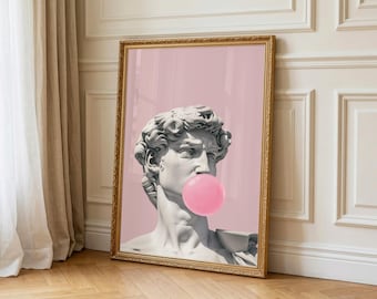 May include: Framed art print depicting a classical bust with a bubblegum bubble. The bust is white, set against a pink backdrop. The gold frame contrasts with the pink and white. The artwork is leaning against a wall and wooden floor.