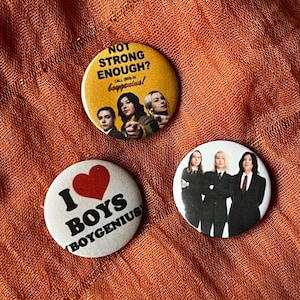 May include: Three round buttons with text and images. One button is yellow with the text "NOT STRONG ENOUGH?" and the words "boygenius!" below an image of three people. Another button is white with a red heart and the words "I BOYS (boygenius)". The last button is white with an image of three people in suits.