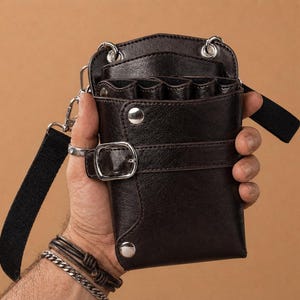 Könnte beinhalten: Dunkelbrauner Friseur-Werkzeug-Organizer aus Leder mit mehreren Taschen und einem Schnallenverschluss. Der Organizer hat einen schwarzen, verstellbaren Riemen und silberfarbene Hardware. Das Design ist kompakt und wirkt langlebig.