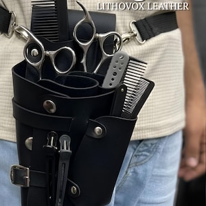 Op de afbeelding: Zwarte leren barbierzak met meerdere compartimenten, met scharen, kammen en andere verzorgingsgereedschappen. De tas wordt om de taille gedragen met een zwarte riem en gesp. Tekst op de afbeelding luidt "Barber Leather Bag" en "LITHOVOX LEATHER".