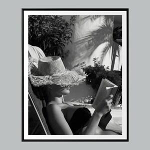 May include: Black and white photograph of a person relaxing outdoors. The person is wearing a straw hat and reading a book while reclining in a chair. The image is framed with a black border.