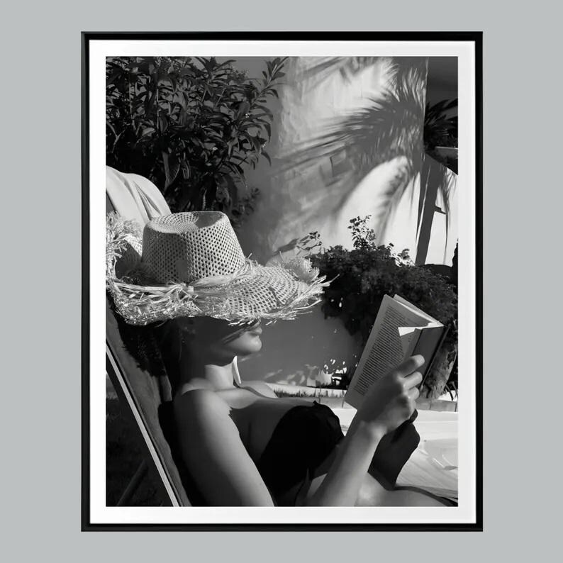 May include: Black and white photograph of a person relaxing outdoors. The person is wearing a straw hat and reading a book while reclining in a chair. The image is framed with a black border.