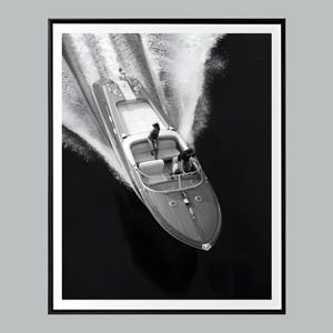 May include: Black and white photograph of a speedboat gliding across dark water, creating a wake. The boat has a wooden deck and seating for passengers. Two people are visible on board. The image is framed in a black border.