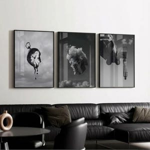 May include: Set of three framed black and white surreal art prints. One print features a melting clock, another shows a car engulfed in smoke, and the third depicts a figure and vehicle. The prints are displayed above a black leather sofa.