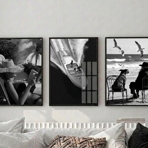 May include: Set of three black and white framed wall art prints. The prints depict a person reading, a speedboat on the water, and a dog on a chair with seagulls flying overhead. The art has a vintage, coastal theme.