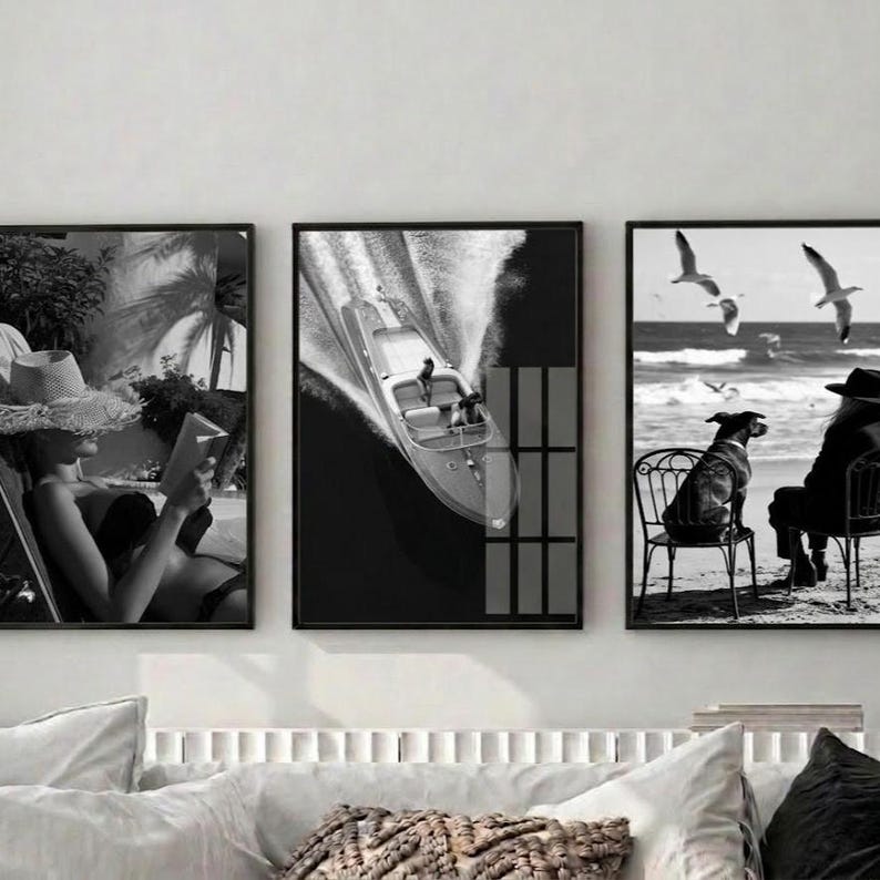 May include: Set of three black and white framed wall art prints. The prints depict a person reading, a speedboat on the water, and a dog on a chair with seagulls flying overhead. The art has a vintage, coastal theme.