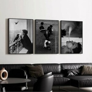 May include: Set of three black and white framed art prints. The prints depict a woman smoking with a crow, people walking on a street, and a person smoking. The art is displayed above a black leather sofa.