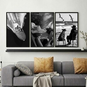 May include: A set of three framed black and white art prints. The prints depict a speedboat on the water, a person reading, and a person with a dog at the beach. The prints are displayed above a gray sofa with gold pillows.
