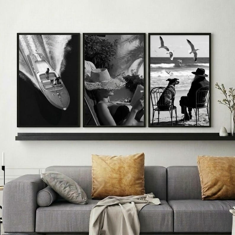 May include: A set of three framed black and white art prints. The prints depict a speedboat on the water, a person reading, and a person with a dog at the beach. The prints are displayed above a gray sofa with gold pillows.