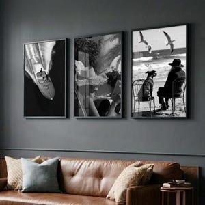 May include: Three framed black and white photographs. The first shows a boat on the water. The second depicts a person reading. The third shows a person and a dog on a beach with seagulls flying overhead. The frames are black.
