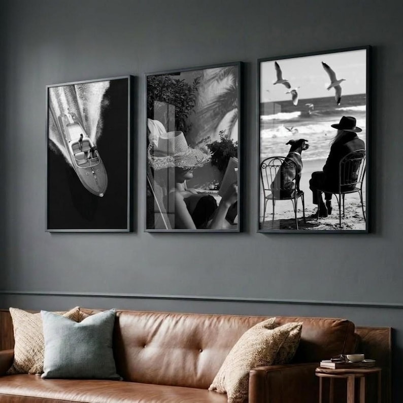 May include: Three framed black and white photographs. The first shows a boat on the water. The second depicts a person reading. The third shows a person and a dog on a beach with seagulls flying overhead. The frames are black.