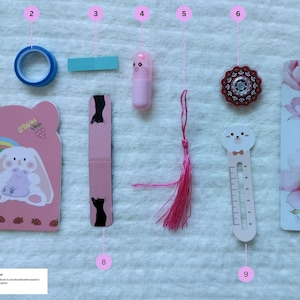 以下が含まれることがあります： ウサギと虹のデザインのピンクのメモ帳、青いマスキングテープ、ピンクのリップクリーム、ピンクのタッセル、装飾ボタン、花の模様のブックマーク、かわいい定規など、文房具のコレクションです。