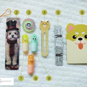 Op de afbeelding: Een verzameling briefpapierartikelen, waaronder een kwast, een hondenbladwijzer, een kleurrijke top, drie capsulevormige pennen met gezichten, een hertenliniaal, een libellenbladwijzer en een notitieblok in de vorm van een hond. De items zijn gerangschikt op een wit oppervlak.