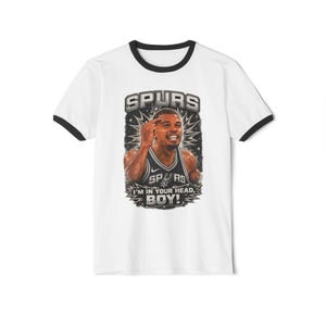 May include: A white ringer t-shirt with black trim on the collar and sleeves. The front features a graphic of a basketball player in a Spurs jersey pointing to their head, with the text SPURS and I'M IN YOUR HEAD, BOY! in a bold font.