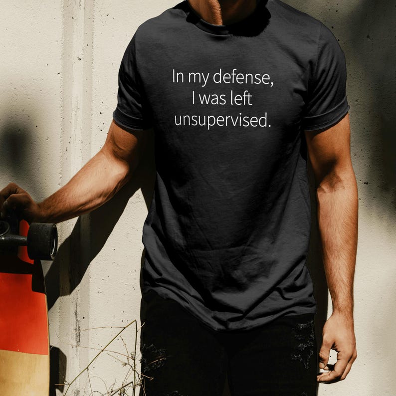 Peut inclure: T-shirt noir avec le texte blanc : &laquo; In my defense, I was left unsupervised. &raquo; Le t-shirt est &agrave; manches courtes et semble &ecirc;tre fait d'une mati&egrave;re douce. La personne tient un skateboard.