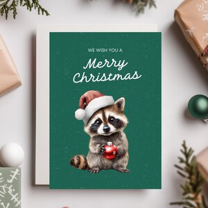 Puede incluir: Tarjeta de Navidad con un mapache con un gorro de Papá Noel y sosteniendo una bola roja. La tarjeta tiene el texto "We wish you a Merry Christmas" sobre un fondo verde. Decoraciones navideñas alrededor de la tarjeta.