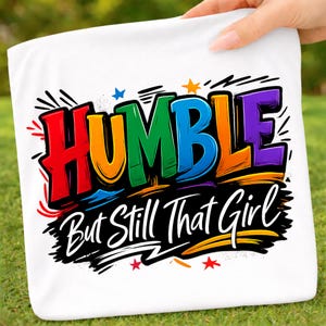 May include: White square towel with the words "HUMBLE" in rainbow colors and "But Still That Girl" in white script. The towel is held up against a green grassy background.