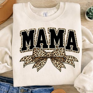 Op de afbeelding: Crèmekleurige sweatshirt met het woord "MAMA" in grote, zwarte blokletters, omlijnd met een luipaardprintpatroon. Een grote luipaardprint strik is gecentreerd eronder. De sweatshirt is een casual, comfortabele stijl.