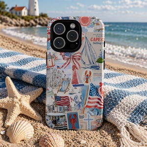 May include: A phone case with a patriotic Cape Cod design featuring a lighthouse, sailboat, American flags, and lobster illustrations. The case is on a striped blue and white beach towel, with seashells and a starfish in the sand.