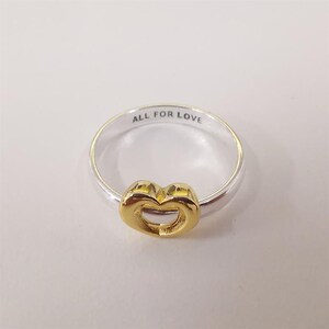 May include: A silver ring featuring a gold heart-shaped design. The ring is inscribed with the words "ALL FOR LOVE". The heart is a bright gold colour.