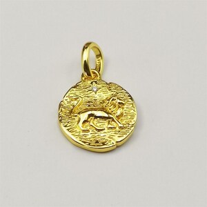 May include: A gold-colored pendant featuring a textured circular design with a lion figure. The pendant has a small loop at the top for a chain and a tiny, sparkling accent. The lion is depicted in a walking pose.