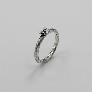 May include: A silver ring featuring a textured band designed to look like tree bark. A small bird-like ornament is perched on the band, with a small gemstone. The ring is a nature-inspired piece of jewellery.