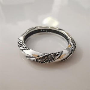 May include: A silver ring with a twisted design. The ring has a band of small, dark-coloured stones along the edges of the twist. The ring is set against a light background.