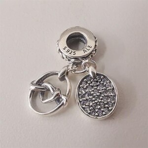 May include: A silver charm with three distinct elements: a knotted heart, a ring, and an oval pendant. The oval pendant is covered in small, clear stones. The charm has the markings "S925 ALE".