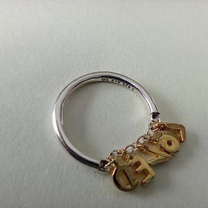 Op de afbeelding: Een zilveren ring met goudkleurige bedels met de tekst "LOVE". De ringband is zilver met de inscriptie "S925 ALE 50". De bedels zijn met kleine kettingen aan de ring bevestigd.