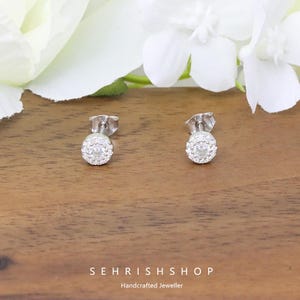 May include: A pair of silver stud earrings with a circular design, each encrusted with small, clear gemstones. The earrings are set against a wooden surface, with white flowers in the background. The text "SEHRISHSHOP Handcrafted Jeweller" is visible.