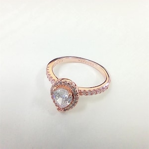 May include: Rose gold ring with a pear-shaped, clear gemstone centre. The ring features a halo of smaller stones around the centre stone and along the band. The brand name "PANDORA" is visible on the ring's band.