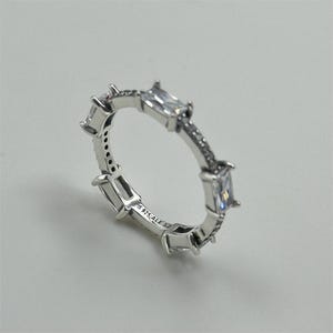 May include: A silver ring with rectangular clear stones and smaller clear stones. The ring is marked with "S 925 ALE 52". The ring is on a white surface.
