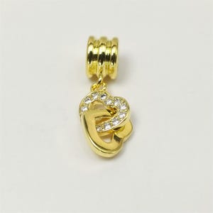 Puede incluir: Un charm dorado con diseño de corazón. El charm presenta dos corazones entrelazados, uno de ellos incrustado con pequeños cristales transparentes. La parte superior tiene un diseño acanalado para sujetarlo a una pulsera o collar.