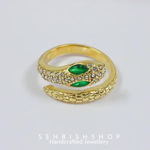 Könnte beinhalten: Ein goldfarbener Schlangenring mit gewundenem Design. Der Schlangenkopf hat zwei smaragdgrüne Augen und ist mit kleinen, klaren Kristallen besetzt. Der Körper des Rings hat ein strukturiertes, schuppenartiges Muster. Der Markenname "SEHRISHSHOP Handcrafted Jewellery" steht unten.