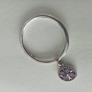 Può includere: Un anello d'argento con un piccolo ciondolo rotondo. Il ciondolo presenta un design a farfalla dettagliato con piccole pietre trasparenti. La fascia dell'anello è liscia e lucida. L'anello è su una superficie bianca.