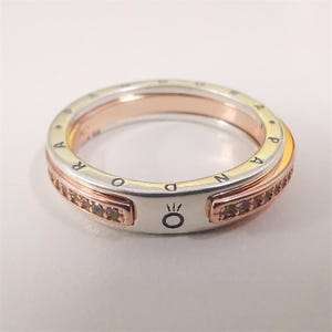 May include: A silver and rose gold ring with a band of small brown gemstones. The ring has the word "PANDORA" engraved around the outer edge. A small symbol is visible on the front of the ring.