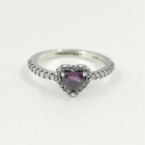May include: A silver ring featuring a heart-shaped, purple gemstone. The gemstone is encircled by a halo of small, clear stones. The band is also set with small, clear stones. The ring is set against a white backdrop.