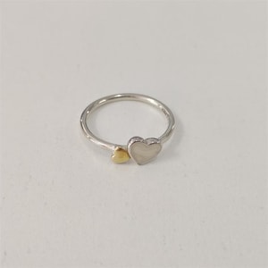 Puede incluir: Un anillo plateado con dos adornos en forma de corazón. Un corazón es dorado y el otro blanco. El anillo está sobre un fondo blanco.