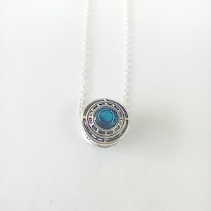 May include: A silver pendant necklace with a circular design. The pendant has a blue centre surrounded by concentric geometric patterns. The necklace chain is delicate and silver, creating a minimalist aesthetic.